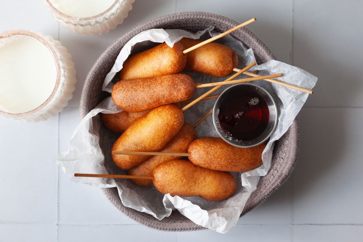 Full yield of Taste of Home Pancakes and Sausage on a Stick in a knit purple gray basket lined with parchment paper and a silver ramekin of pancake syrup, glasses of milk