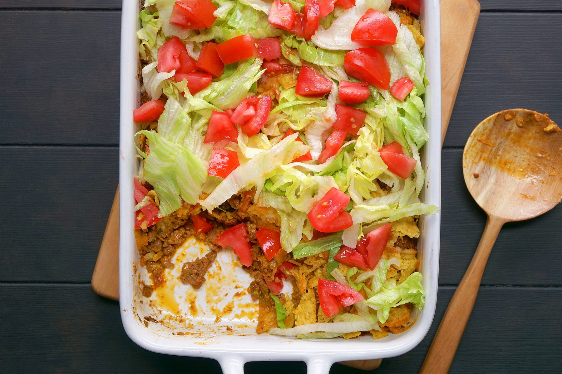 overhead shot; black background; Taco Casserole in a long baking dish placed over wooden board with woodeb serving spoon;