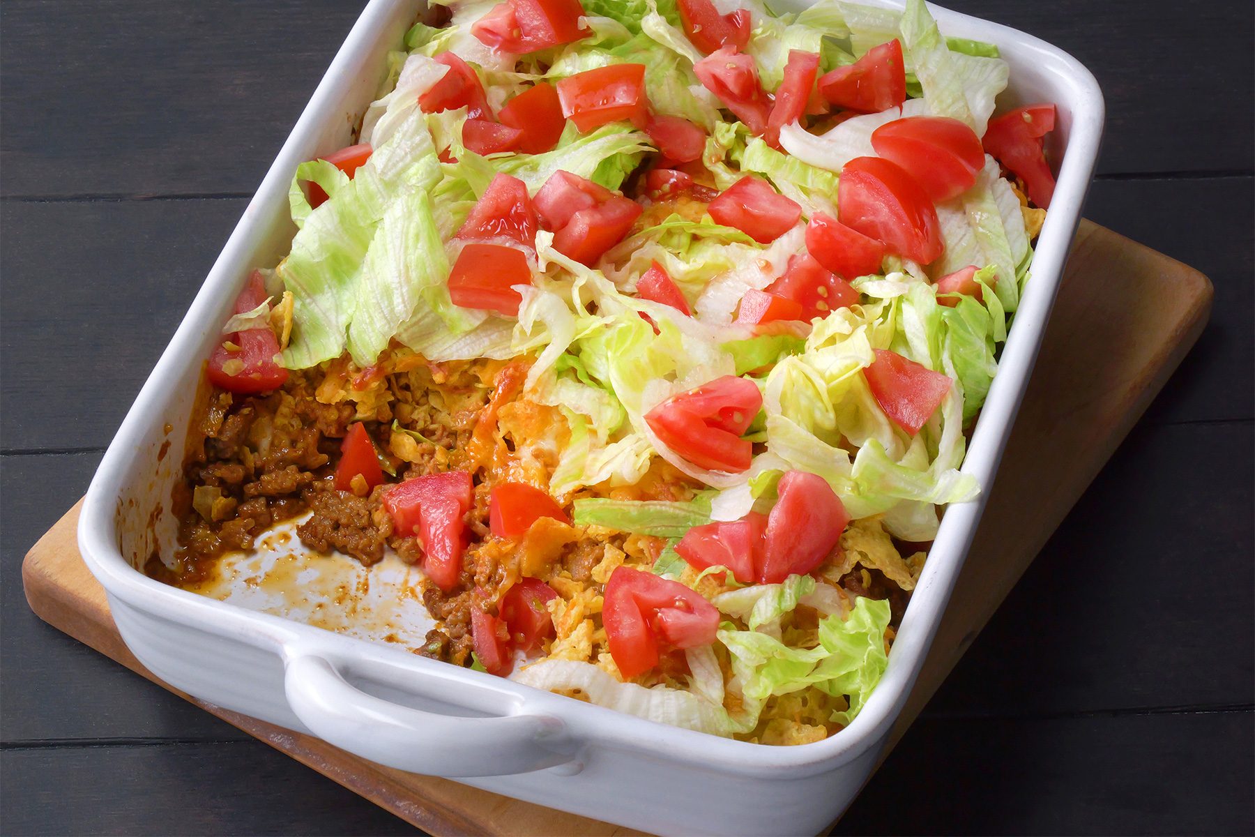 3/4th shot; black background; Taco Casserole in a long baking dish placed over wooden board with woodeb serving spoon;