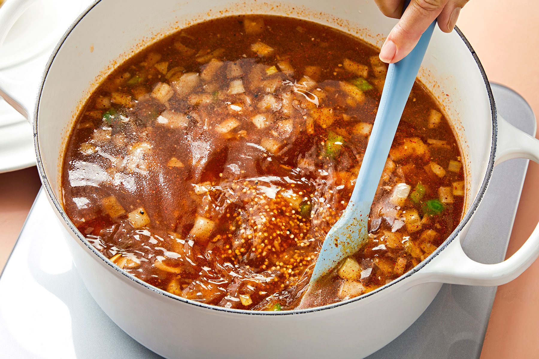 A person stirs a pot of mixed vegetable soup with chopped onions, green peppers, and various spices. The broth is a rich, dark, reddish-brown color, and the pot is white and placed on a stovetop.