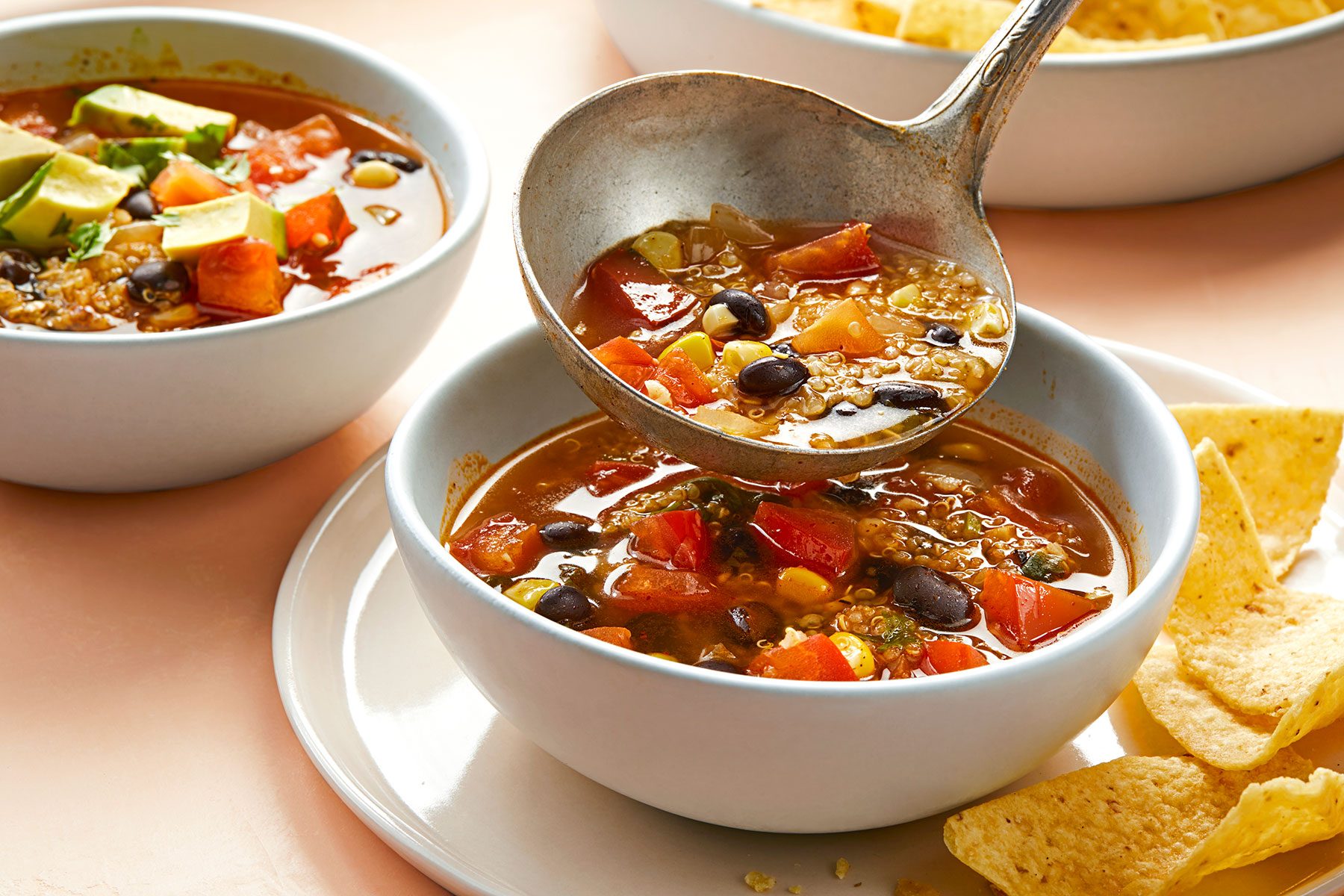 A close-up of three bowls of a hearty soup filled with vegetables, beans, and grains. One bowl is being served with a ladle. Tortilla chips are on the side of one bowl. The soup is garnished with avocado and is set on a light-colored table.