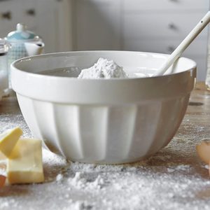 A white mixing bowl filled with flour, a spoon resting inside it. The surrounding surface is dusted with extra flour and there are broken eggshells and pieces of butter nearby, suggesting ongoing baking preparations in a kitchen.