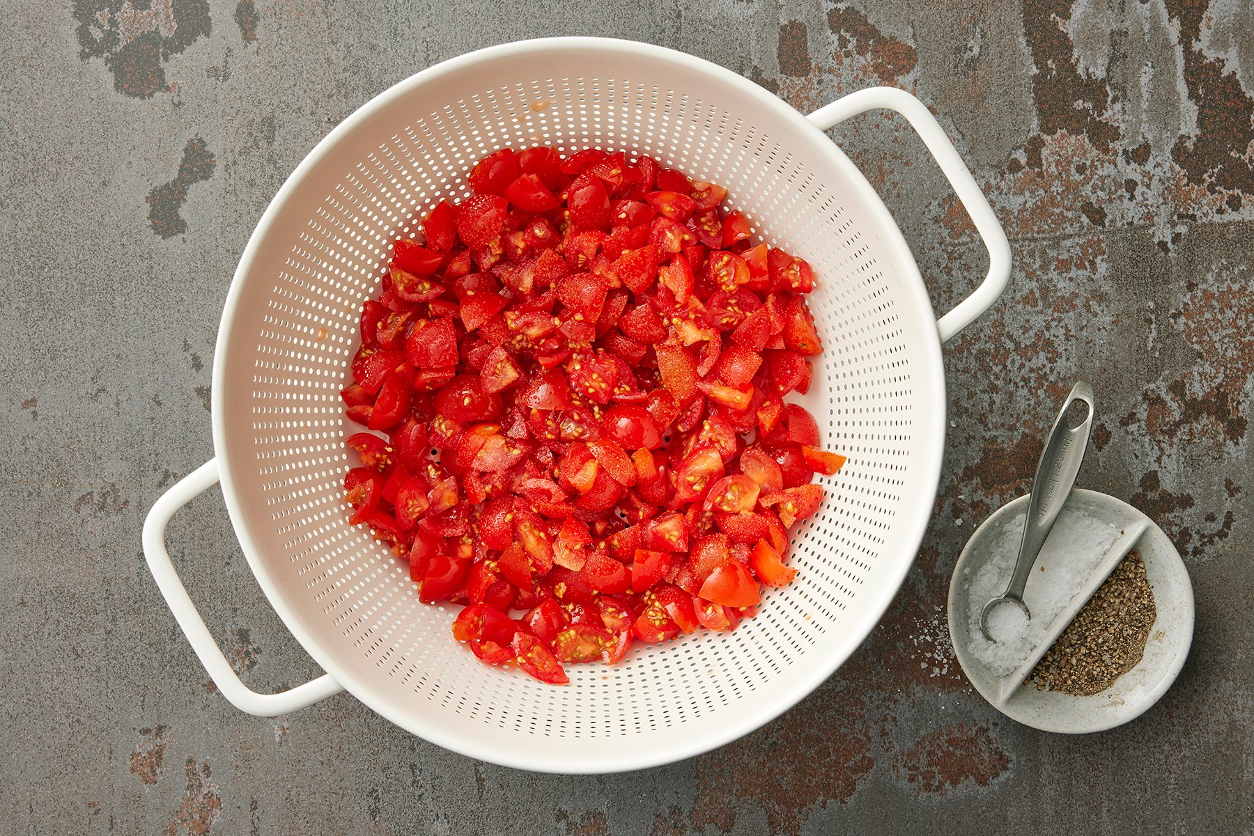 A white colander filled with freshly diced tomatoes sits on a gray, textured surface. Next to the colander, there is a small ceramic dish containing ground pepper and a small spoon.