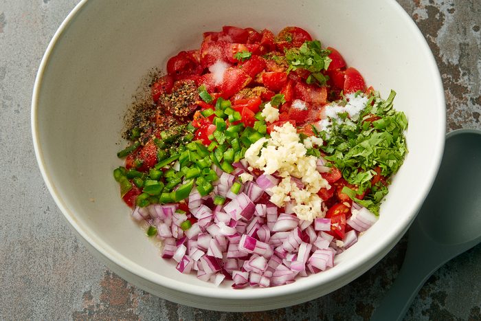 A large white bowl contains chopped red onions, minced garlic, diced green bell peppers, chopped tomatoes, chopped cilantro, and assorted spices. A grey serving spoon is placed beside the bowl on a textured surface.