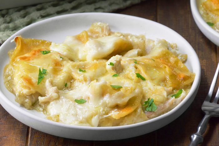 3/4th shot; wooden background; A plate of Chicken Enchilada Bake is showcased, featuring a generous portion of the dish in a white plate, Layers of tortillas, chicken, cheese, and a creamy sauce are visible, The enchiladas are topped with a sprinkle of fresh parsley