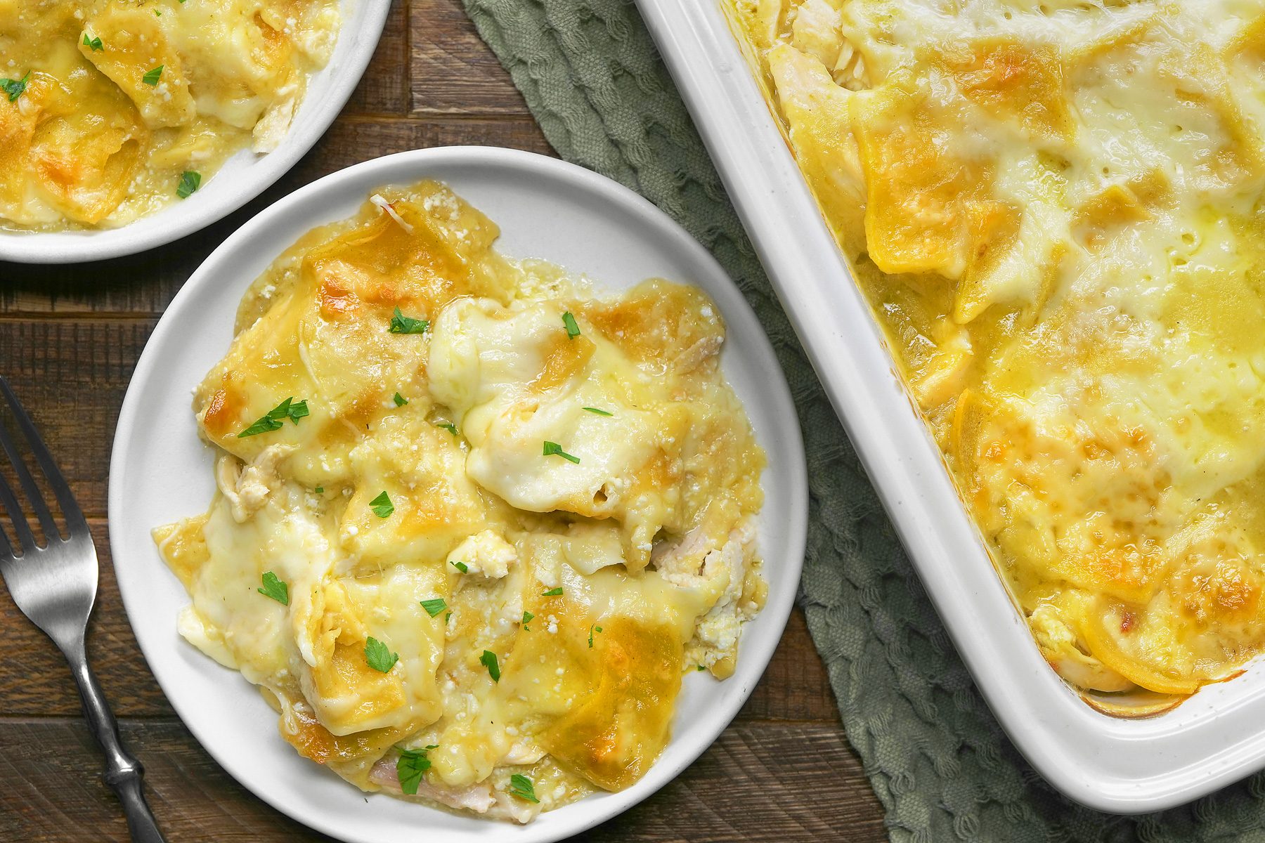 overhead shot; wooden background; Chicken Enchilada Bake is showcased, featuring layers of tortillas, chicken, cheese, and a creamy sauce, The dish is bubbling with melted cheese and has a golden-brown crust, A single serving is plated alongside the main dish, revealing the creamy filling and the melted cheese, Fresh parsley is sprinkled on top for added flavor and color, a sliver fork is also visible