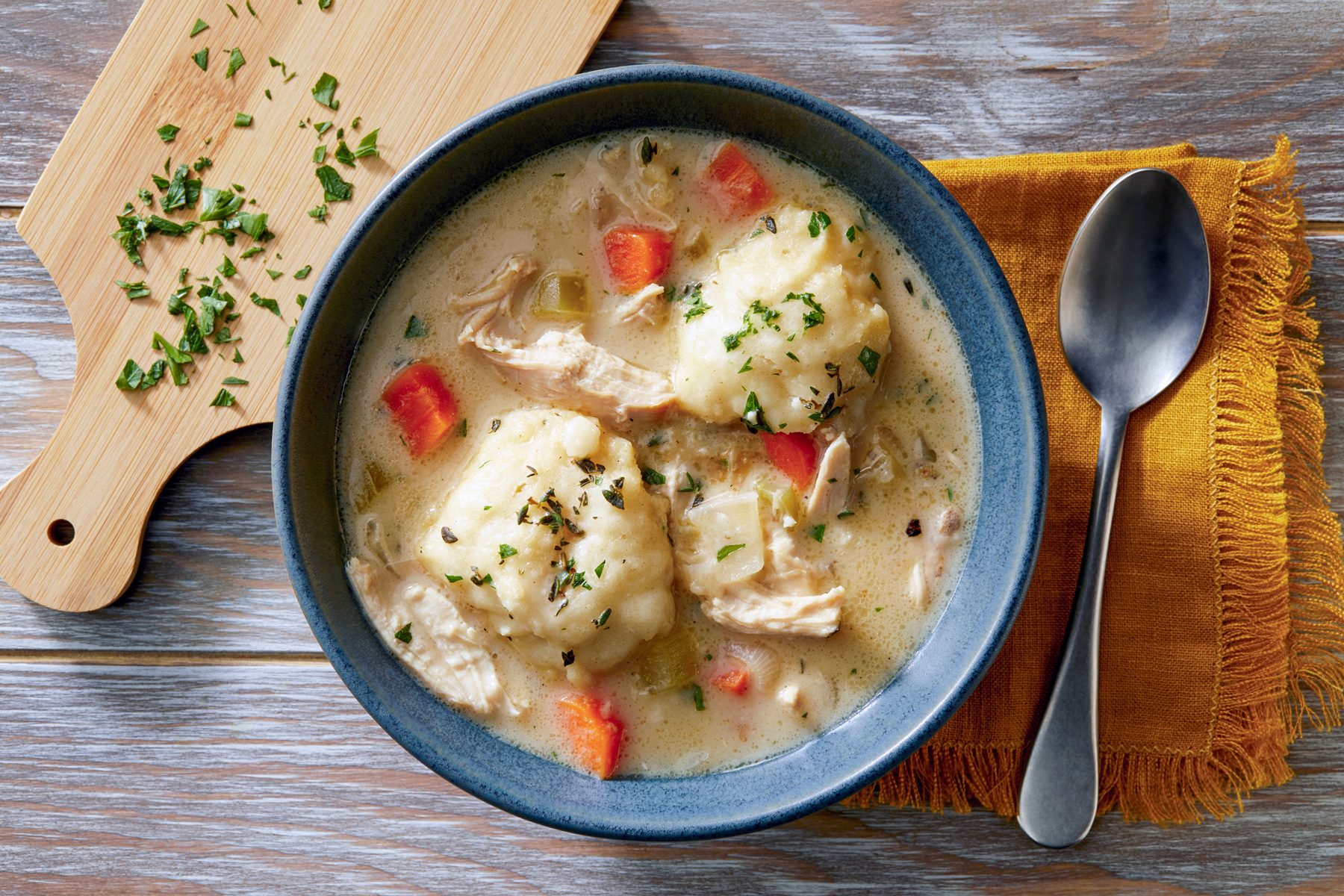 A bowl of Chicken and Dumplings is the centerpiece of the image, The bowl is placed on a wooden table, and a spoon and a cutting board are visible in the background.
