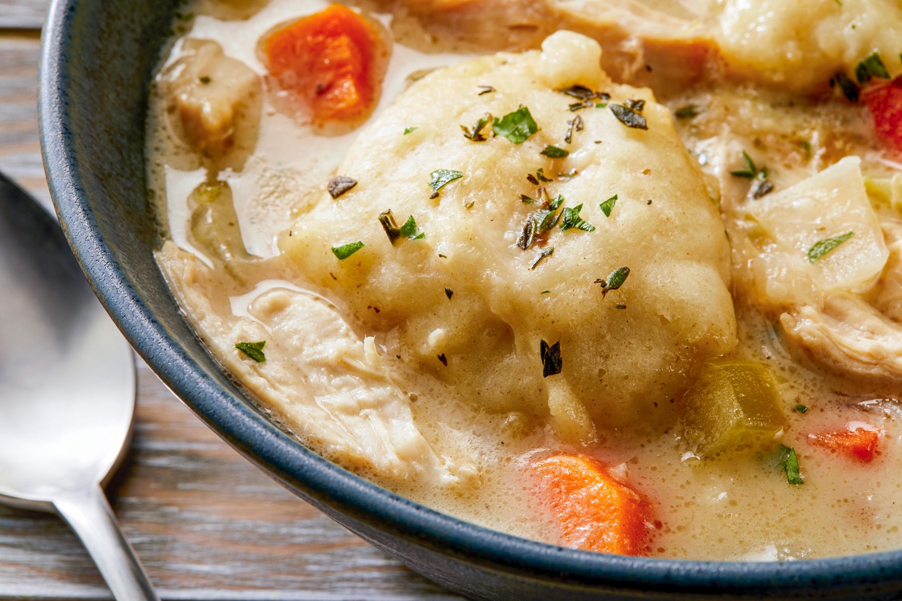 a close-up of a bowl of Chicken and Dumplings, The soup is a creamy white color and is filled with pieces of chicken, vegetables, and soft, fluffy dumplings, a spoon is place aside