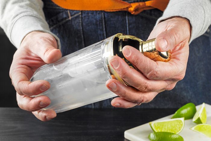 Eye angle view shot of shaker; cover with lid and shake for 10-15 seconds; or until condensation forms on outside of shaker; lime wedges; chopping board; black wooden base