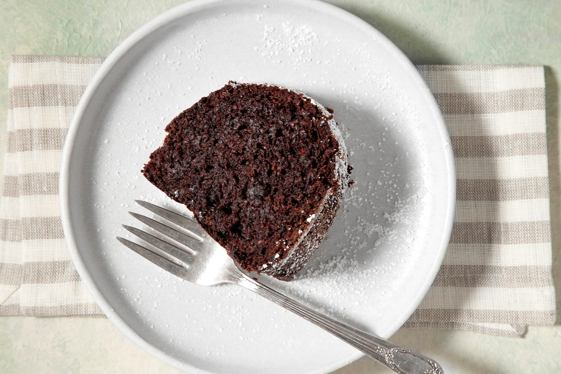 overhead shot; light green background; A slice of chocolate cake on a white plate; It's dusted with powdered sugar; silver fork placed on white plate over kitchen towel