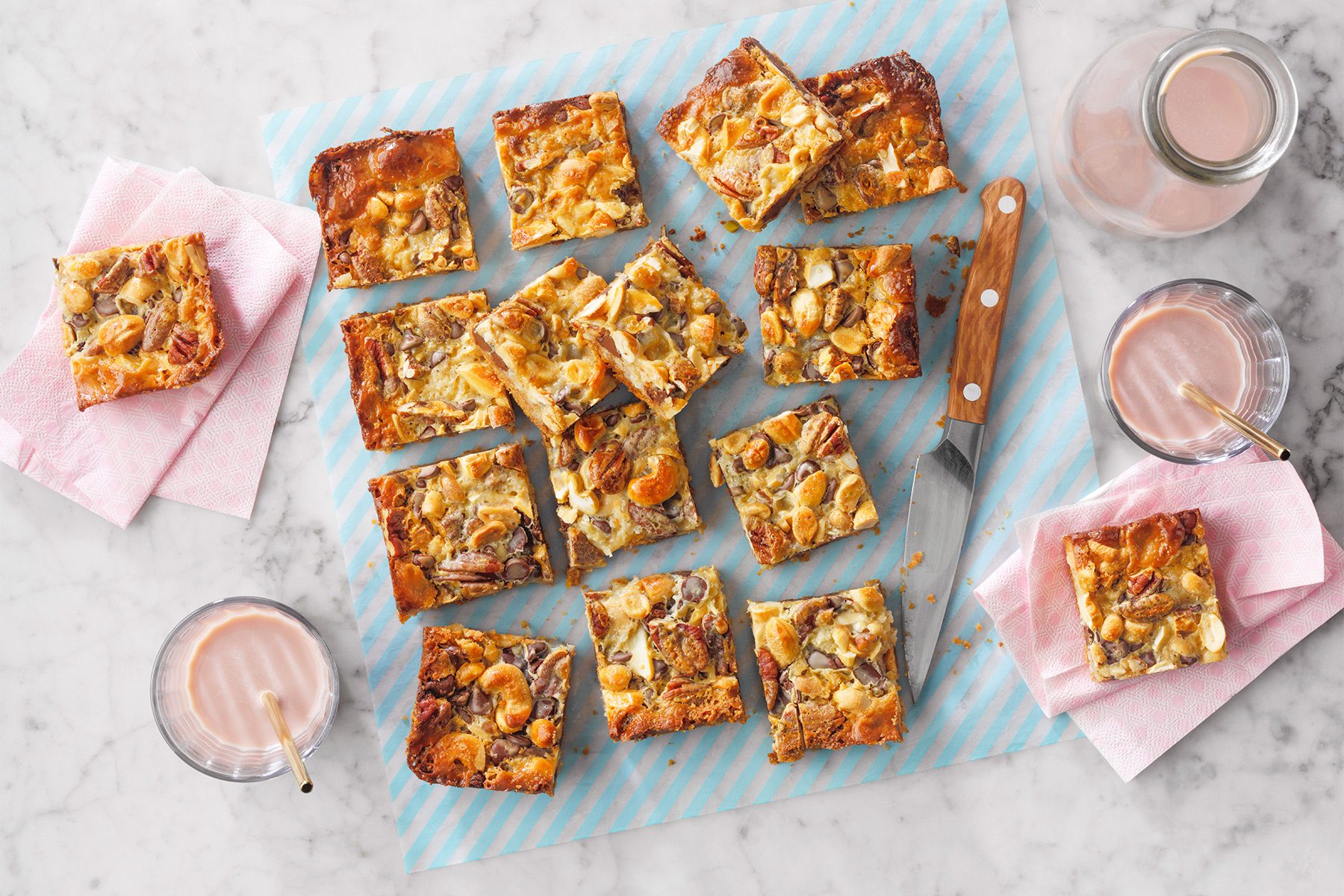 A marble countertop features a blue-striped baking sheet with several pieces of nut and dried fruit-topped bar cookies. Two glasses of chocolate milk with straws and a knife are nearby, along with napkins holding additional cookie pieces.