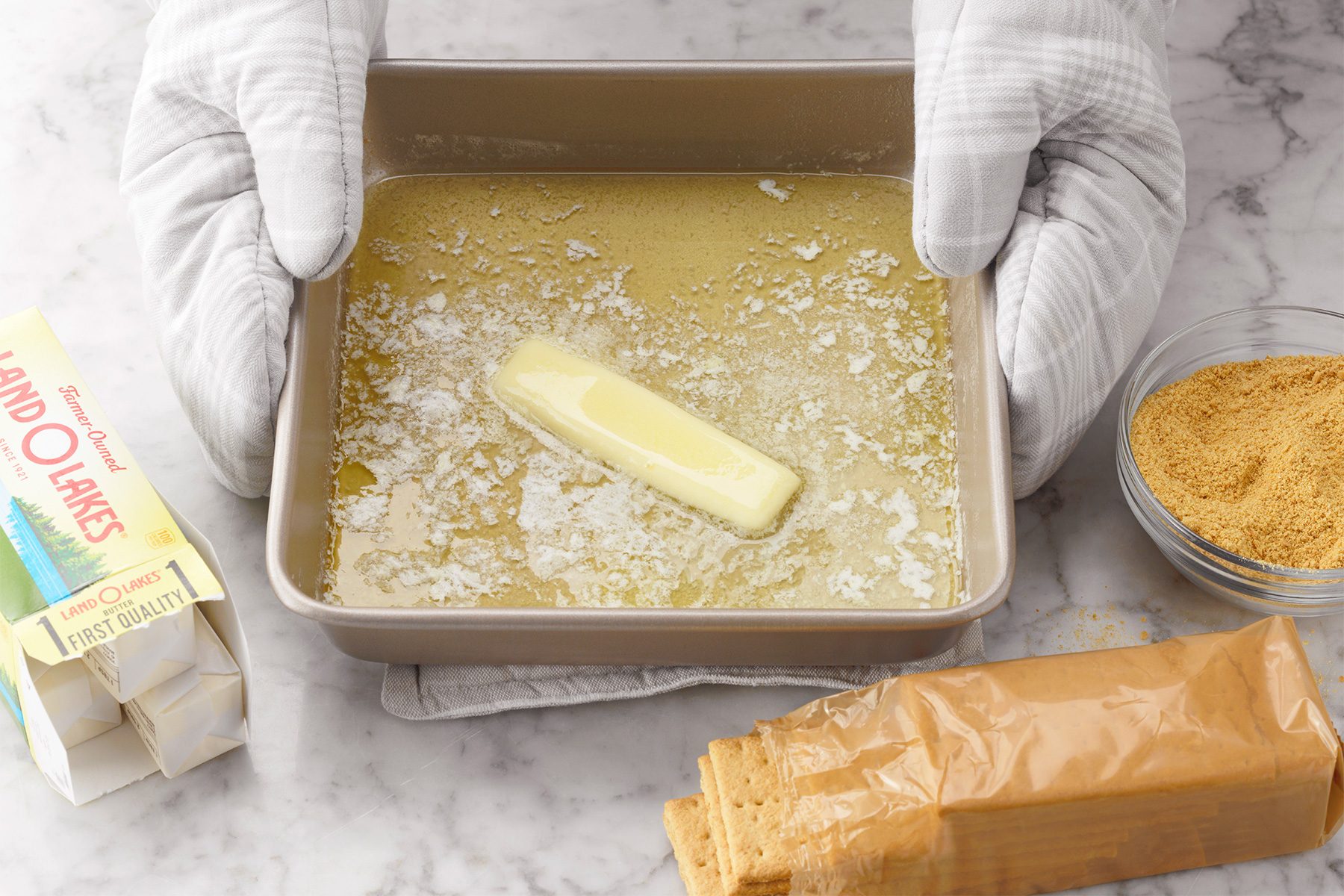 A person wearing oven mitts holds a square baking pan with melted butter. Surrounding the pan are a pack of graham crackers, a partially opened sleeve of wax paper, and a bowl of crushed graham cracker crumbs on a marble surface.
