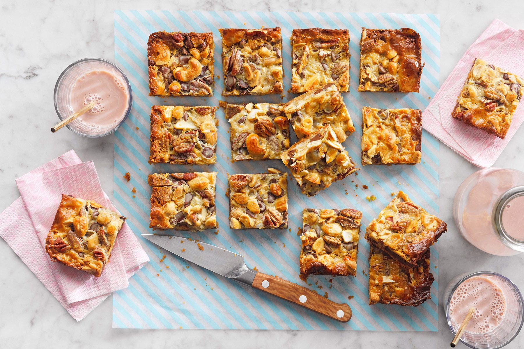 Squares of fruit and nut blondies are arranged on a light blue striped surface. A wooden-handled knife is positioned among the blondies. Two glasses of pink beverage with straws and pink cloth napkins are placed around the blondies on a marble background.