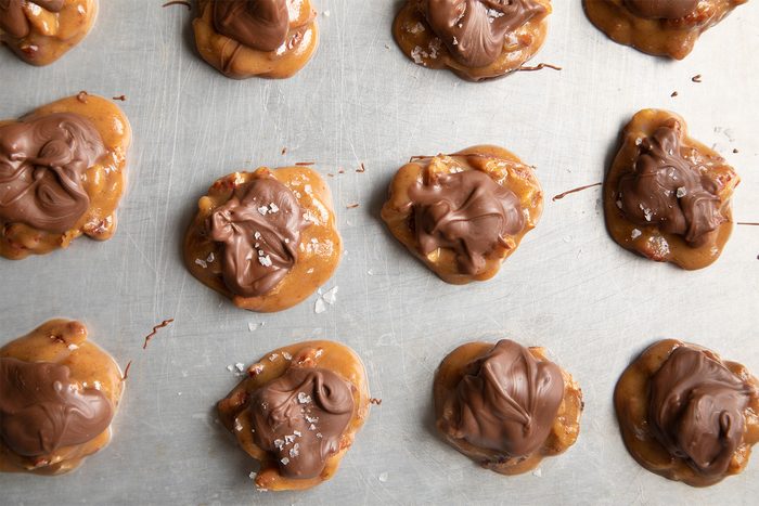 Several clusters of chocolate-coated caramel pecan candies are spaced out on a metal baking sheet. Each cluster appears to be handmade, with a slightly irregular shape and visible pecan pieces beneath the smooth chocolate and caramel coating.