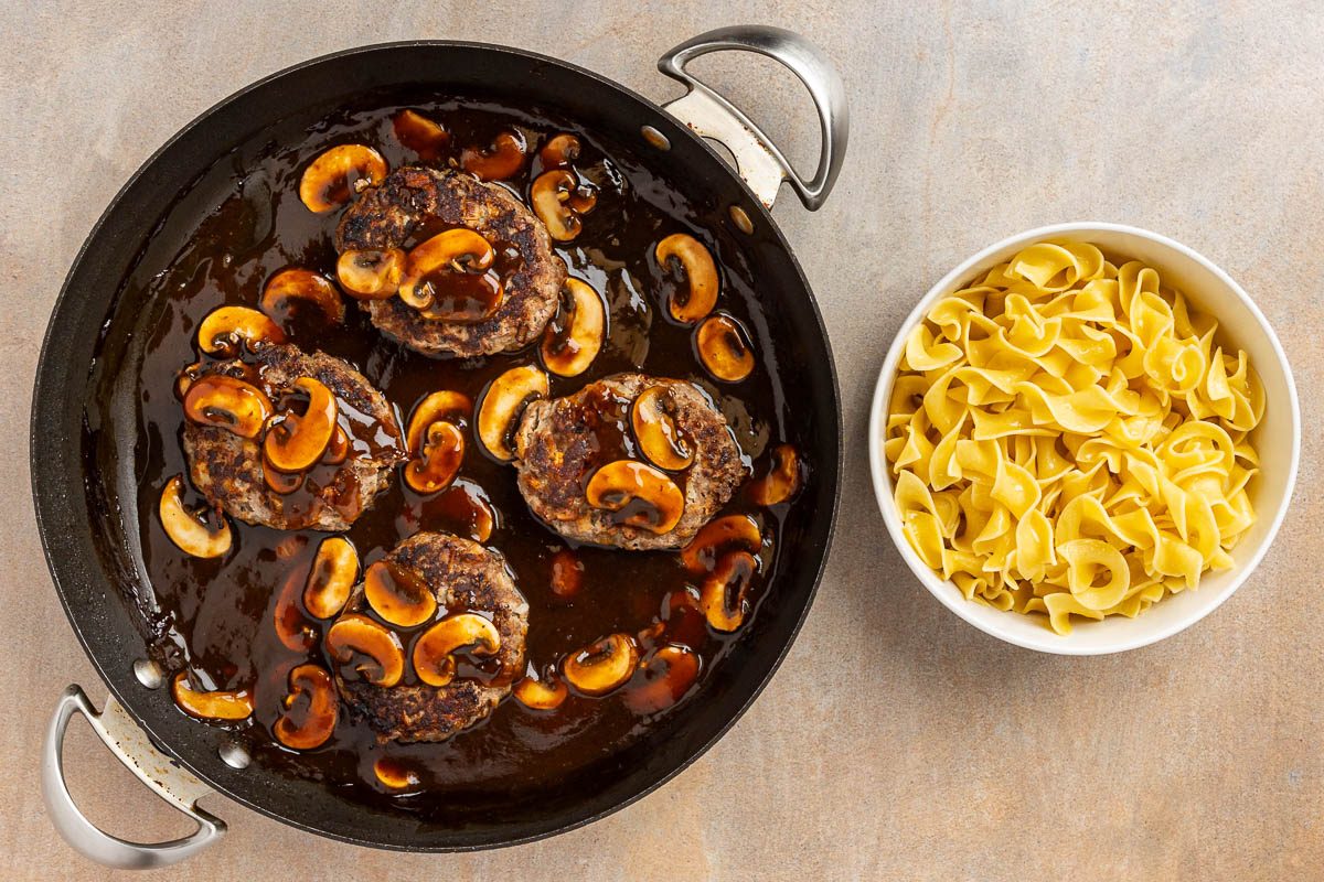 Old Fashioned Salisbury Steak in a pan with a bowl of noodles to the side