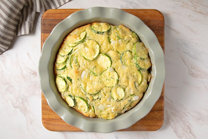 A zucchini casserole sits in a round, fluted dish atop a wooden board on a marble countertop. The surface of the casserole is golden and speckled with visible zucchini slices. A striped cloth is partially visible in the top left corner.