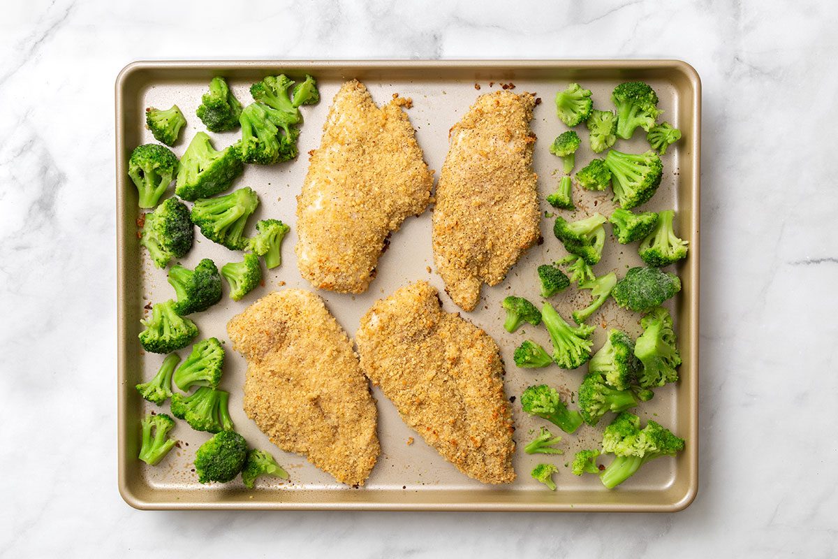 Crumb-coated chicken breasts on a baking sheet with broccoli florets for step two of Baked Chicken Parmesan recipe for Taste of Home