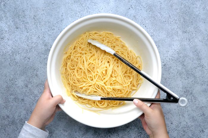 Hands holding a white bowl filled with cooked spaghetti, using tongs for serving. The background is a textured gray surface.