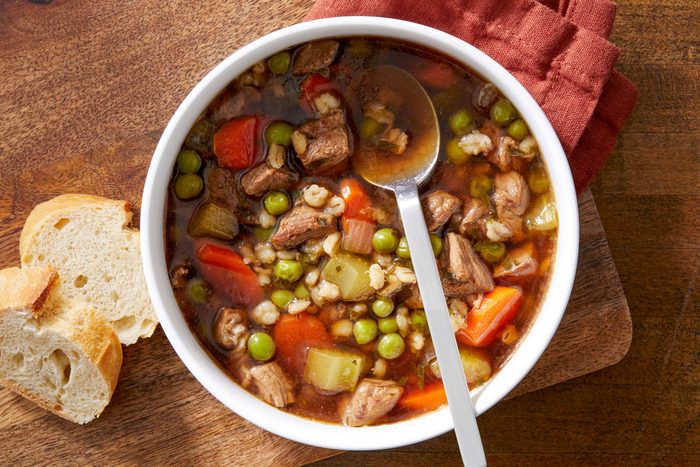 A hearty beef and barley soup is served in a bowl with a spoon and crusty bread on the side