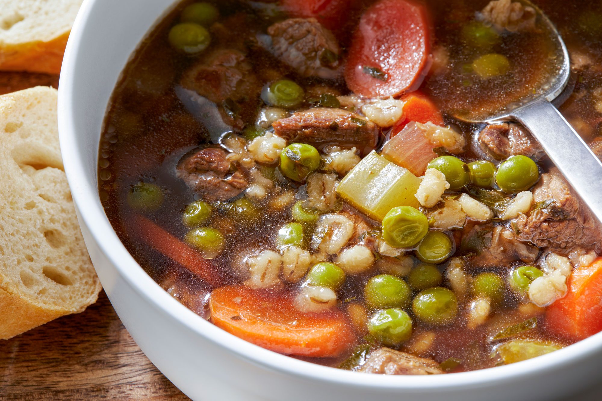 A hearty beef and barley soup is served in a white bowl with a spoon and crusty bread on the side