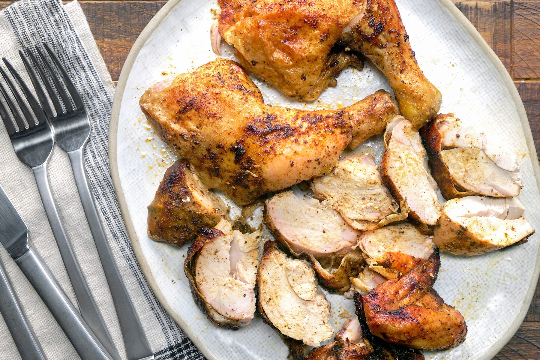 A plate of roasted chicken, including whole legs and sliced breast pieces, is served. The chicken is seasoned and arranged on a white plate, next to a cloth napkin and several forks on a wooden table.