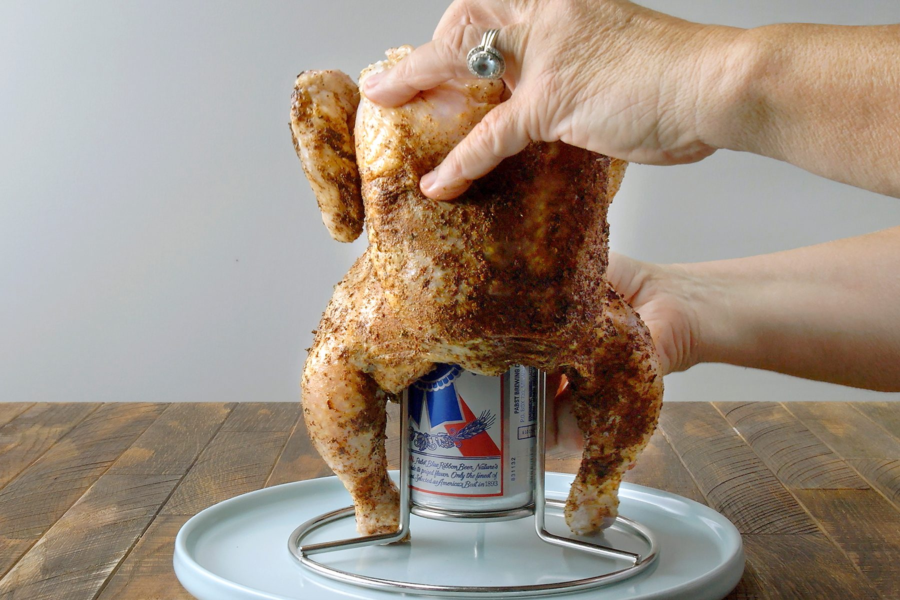 A person's hands preparing beer can chicken; a whole seasoned chicken is placed on a can of beer on a metal stand on a round plate. The wooden surface and plain background are visible.