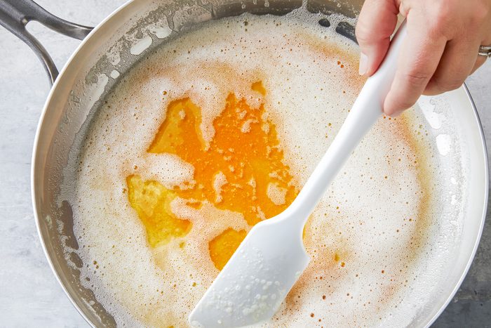 A small saucepan on a stovetop with melting butter, bubbling and turning golden brown. A wooden spoon is stirring the butter occasionally, with a timer or clock showing a duration of 5 to 7 minutes nearby.