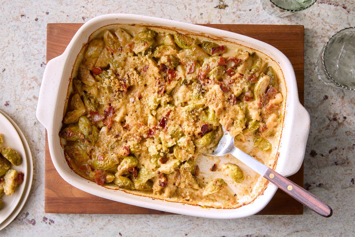 Overhead shot of Brussels Sprouts Casserole; in a baking pan; on wooden board; serving spoon; serve on plate; water glasses; marble surface;