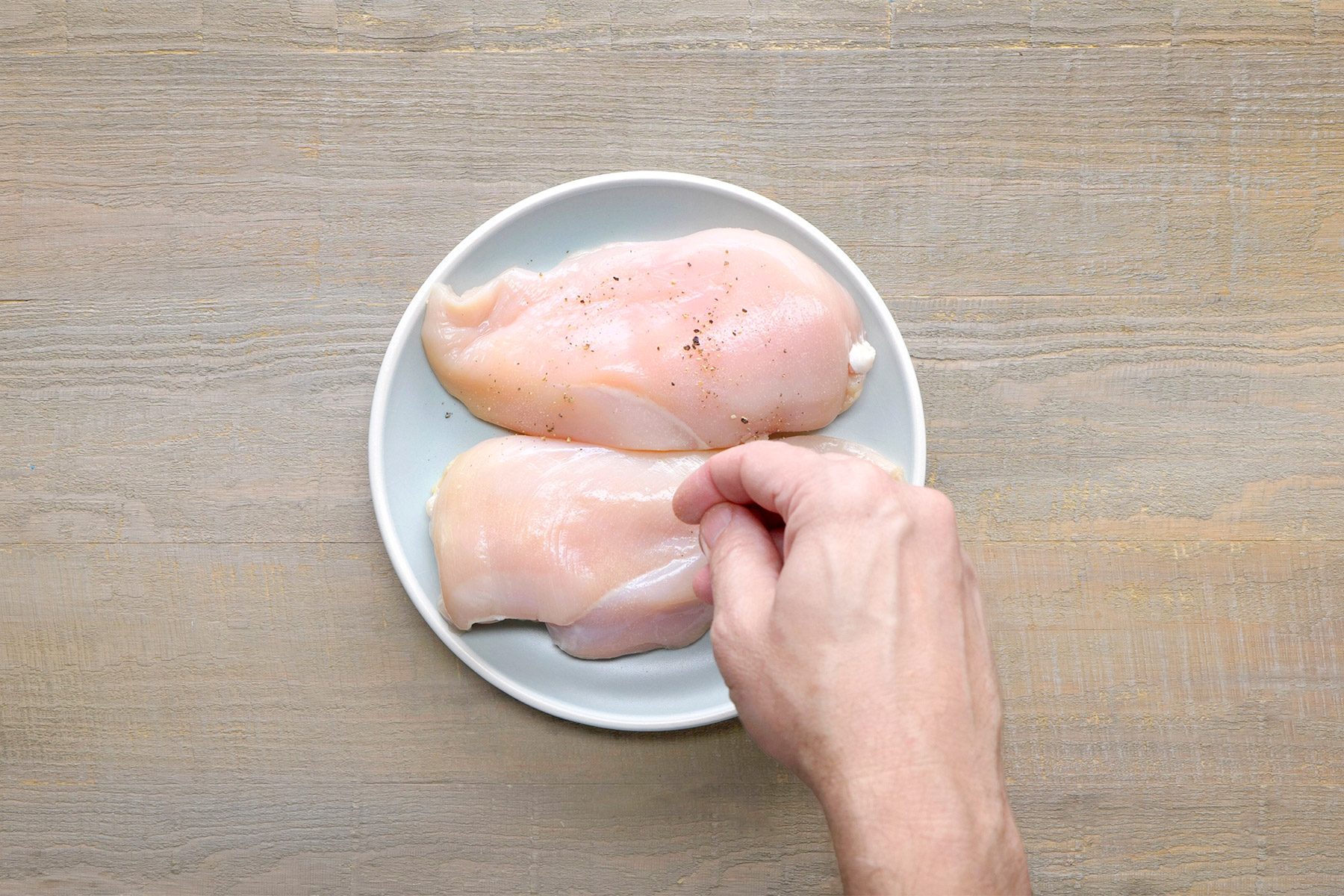 overhead shot of two raw chicken breasts placed on a white plate, The chicken breasts are a pale white color and have a slightly moist texture. A person's hand is touching one of the chicken breasts, The chicken breasts are seasoned with a sprinkle of black pepper;