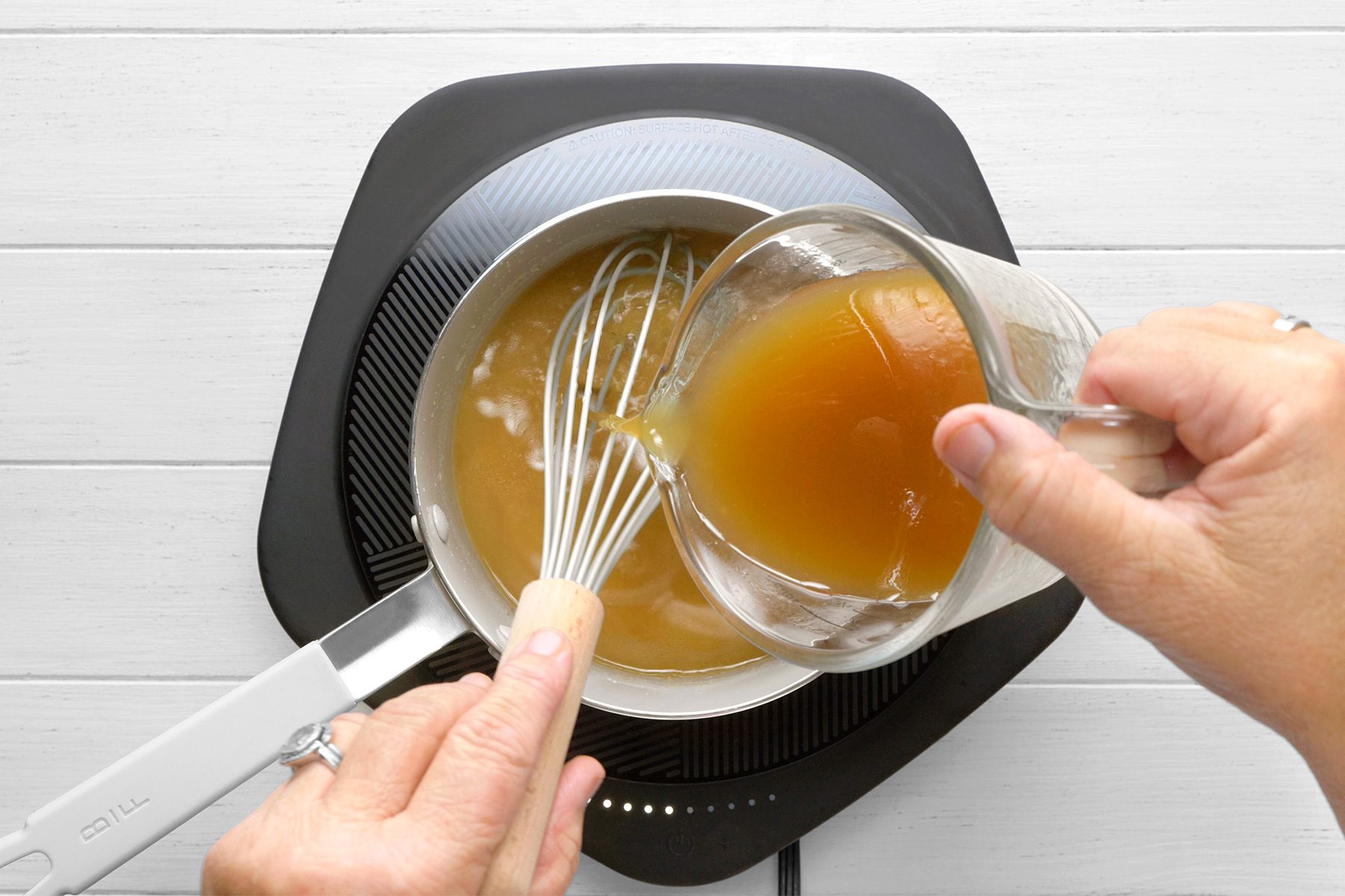 overhead shot of a person pouring a liquid from a measuring cup into a saucepan, The liquid is a dark brown color and has a thick consistency, The saucepan is on an electric induction cooktop and contains a mixture of butter and flour, The person is holding a whisk in the saucepan;