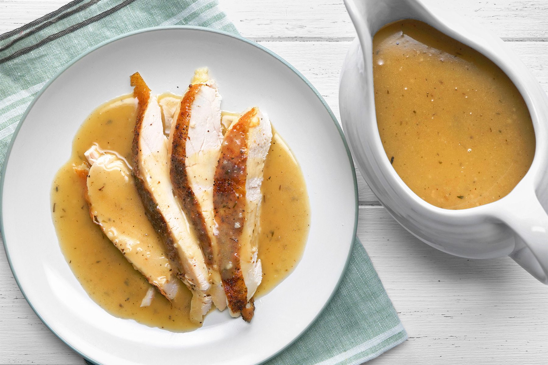 overhead shot of a delicious dish of chicken gravy, The chicken is sliced and arranged neatly on a white plate, showcasing a golden-brown seared crust, Beside the plate, there’s a gravy boat filled with creamy, herb-speckled gravy, The dish is presented on a white wooden table with a folded green-striped napkin under the plate;