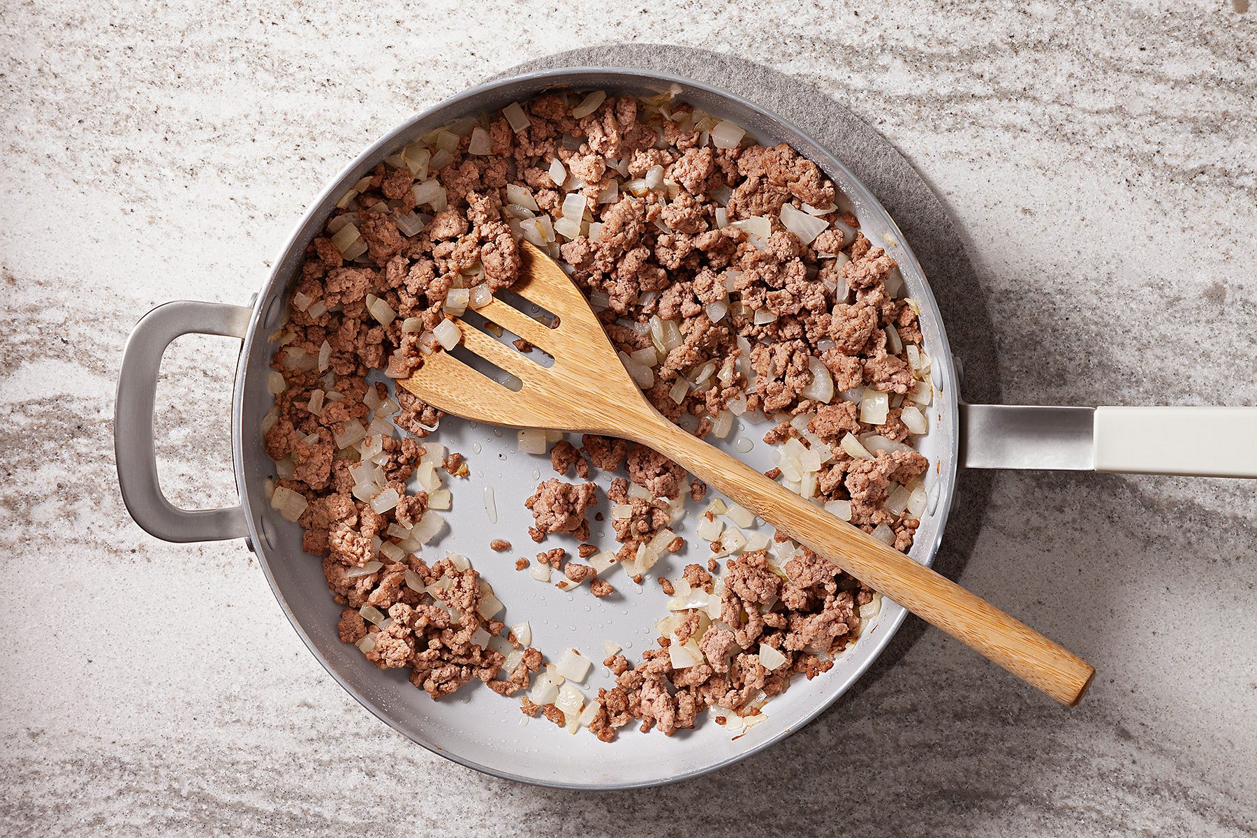 A frying pan with cooked ground beef and chopped onions, stirred with a wooden spatula, placed on a marbled countertop.