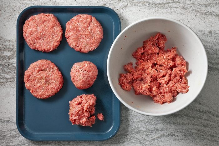 Ground meat is being prepared on a kitchen counter. Five formed patties are arranged on a blue tray to the left, while a white bowl on the right contains more raw meat ready to be shaped. The surface is a light gray textured countertop.