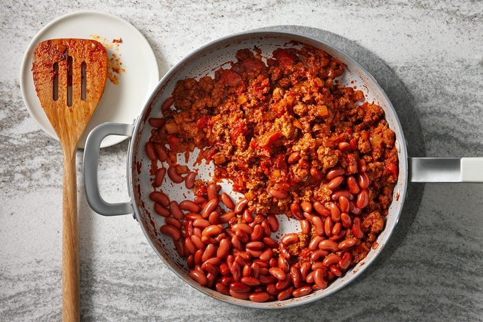 A skillet containing cooked ground meat and kidney beans in a red sauce sits on a gray counter. A wooden spatula with sauce rests on a small white plate next to the skillet, with remnants of sauce visible on it.