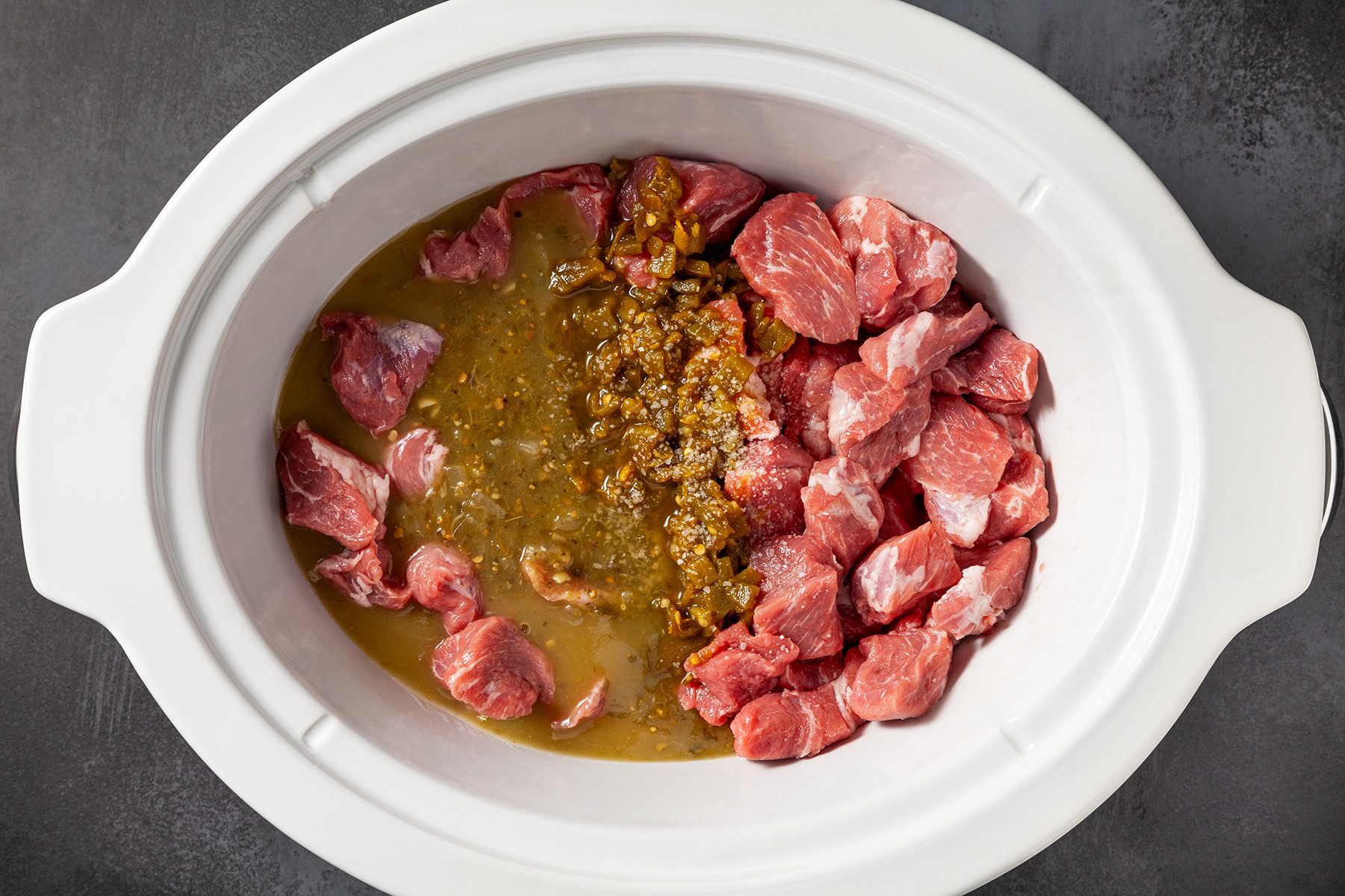 Chunks of raw meat and a mixture of green chile sauce are placed side by side in a white slow cooker.