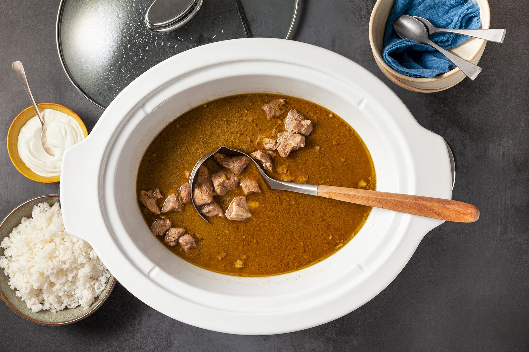 A white slow cooker filled with curry and chunks of meat, with a wooden ladle resting inside. Surrounding it are bowls containing steamed white rice, sour cream, a spoon, and a napkin. A lid is partially shown on the side.
