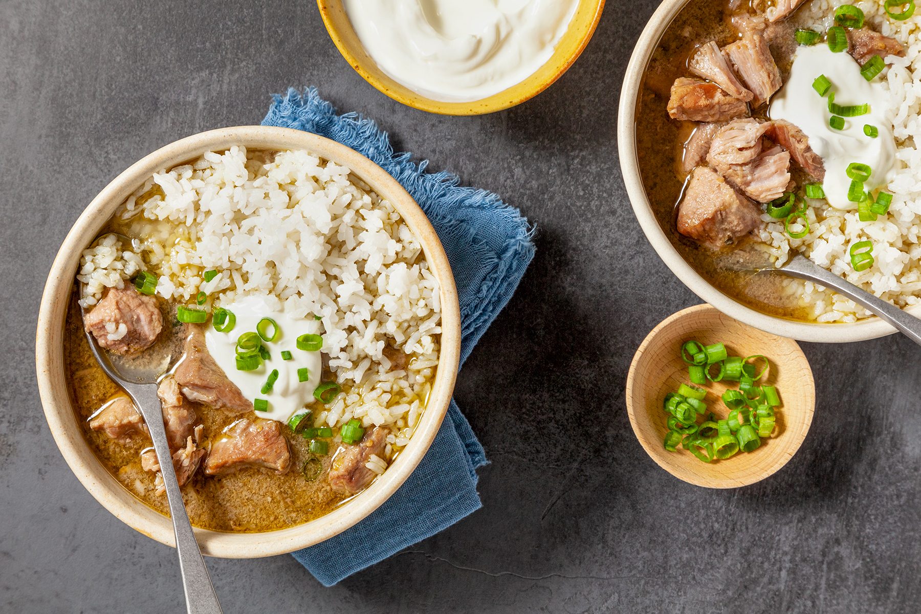 Two bowls of rice topped with chunks of meat, a dollop of sour cream, and chopped green onions. A small wooden bowl with green onions and a yellow bowl of sour cream are nearby, set on a dark textured surface with a blue cloth.