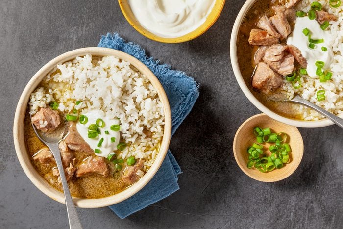 Two bowls of rice topped with chunks of meat, a dollop of sour cream, and chopped green onions. A small wooden bowl with green onions and a yellow bowl of sour cream are nearby, set on a dark textured surface with a blue cloth.
