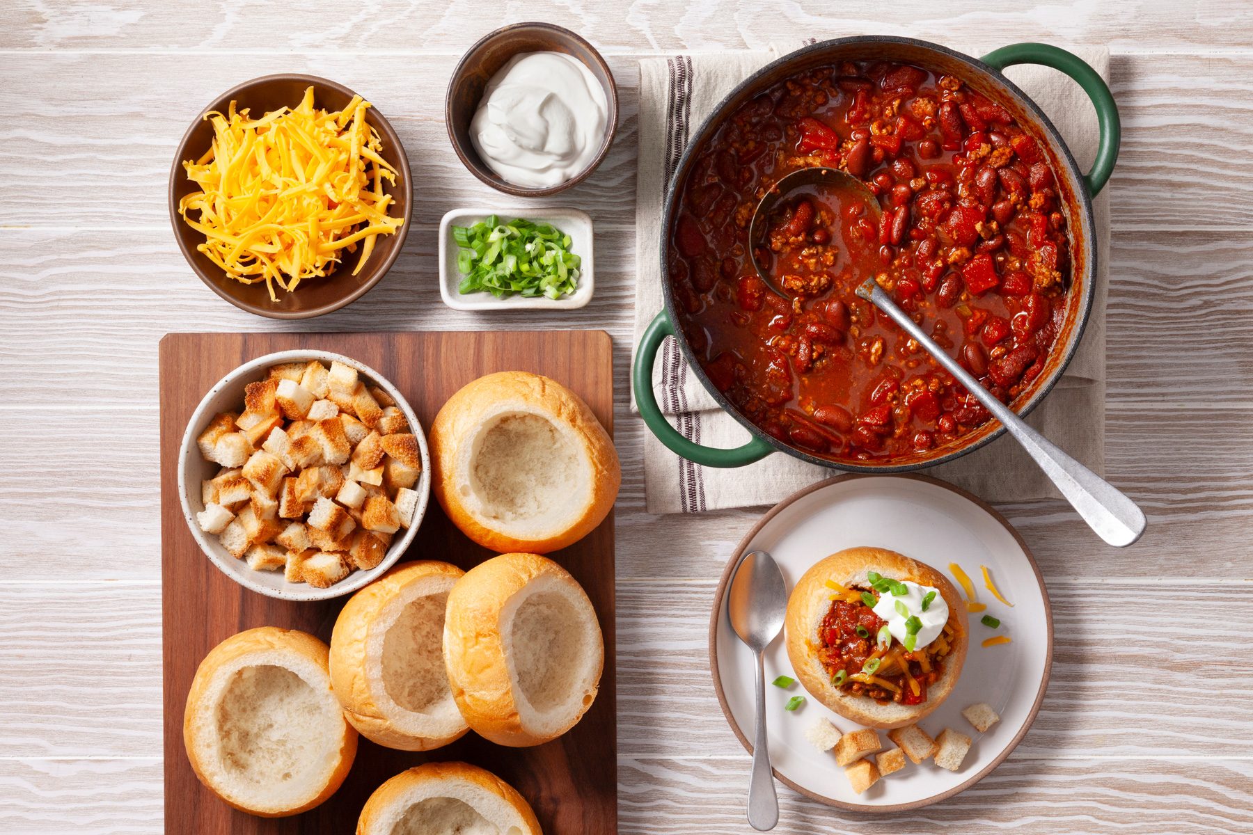 An overhead shot of a delicious and hearty chili dish, perfect for a cozy meal. A large green Dutch oven filled with thick, red chili serves as the centerpiece of the image. The chili is loaded with ground beef, beans, and vegetables, garnished with shredded cheese and chopped green onions. Surrounding the chili are several hollowed-out bread bowls, ready to be filled with the flavorful chili. Additional ingredients, such as shredded cheese, sour cream, and croutons, are also present.