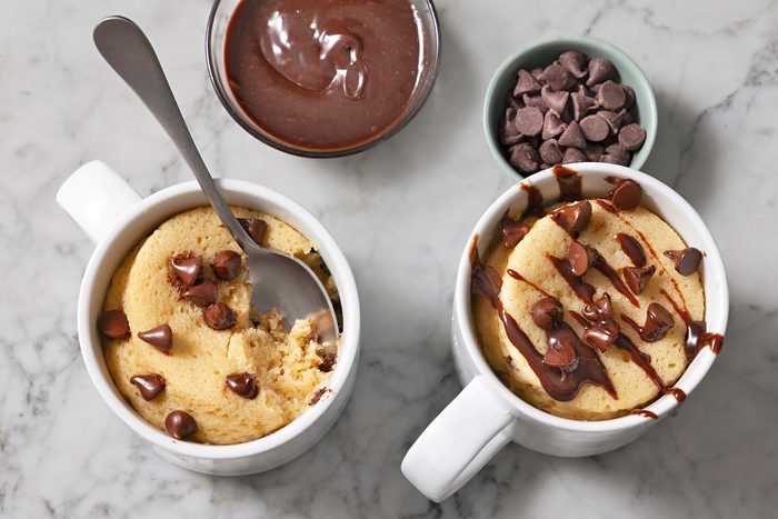 Two chocolate Chip Mug Cakes. One mug has a spoon in it. Melted chocolate and chocolate chips can be seen next to them.
