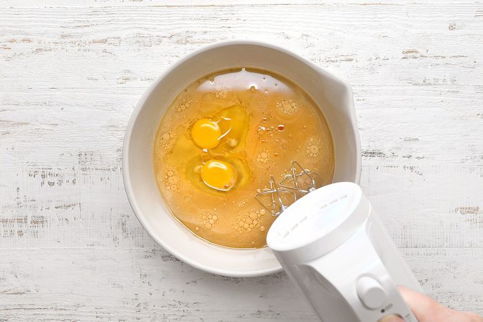 A top-down view of a white mixing bowl containing two cracked eggs and other ingredients. A white electric hand mixer is partially visible on the right, hovering over the bowl, ready to mix the contents. The background is a white wooden surface.