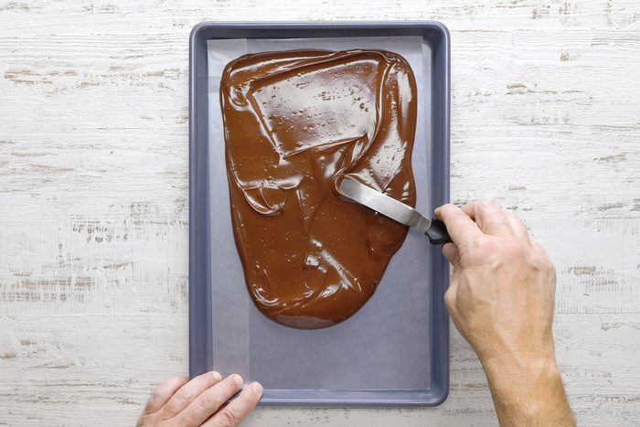 Hands spreading melted chocolate evenly on a baking tray using a spatula, set on a light wooden surface.