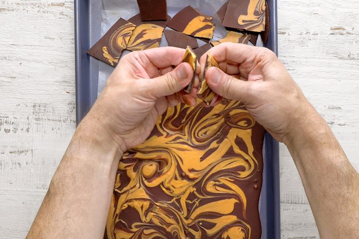 Hands breaking a chocolate bark with a marbled pattern of dark and caramel colors, placed on a baking sheet.