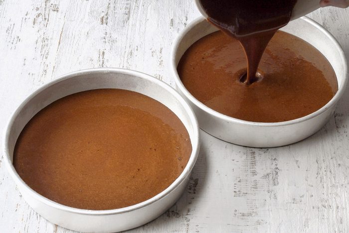 Two round cake pans filled with chocolate batter sit on a white wooden surface. A hand is pouring more batter into one of the pans from a white bowl.