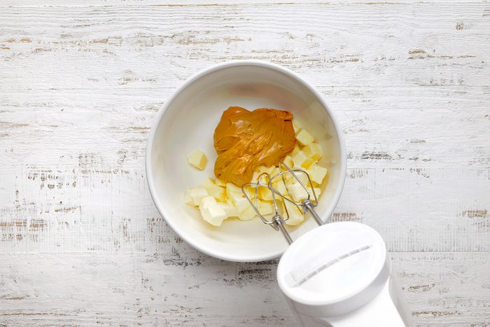 A white mixing bowl contains chunks of butter and a dollop of peanut butter. A hand mixer with beaters is ready to blend the ingredients. The setup is on a rustic white wooden surface.