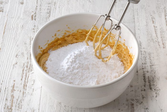 A mixing bowl on a white wooden surface contains peanut butter and powdered sugar, with an electric mixer resting on the edge, partially immersed in the ingredients.