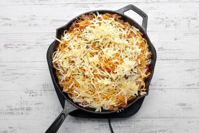 A skillet filled with bacon, beans, sauces, spices, spaghetti and topped with cheese