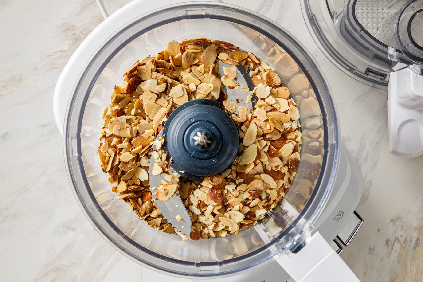 A food processor with a clear bowl containing a generous amount of raw almonds. The processor is plugged in, ready for use, with the lid slightly ajar.