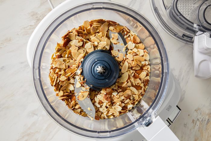 A food processor with a clear bowl containing a generous amount of raw almonds. The processor is plugged in, ready for use, with the lid slightly ajar.