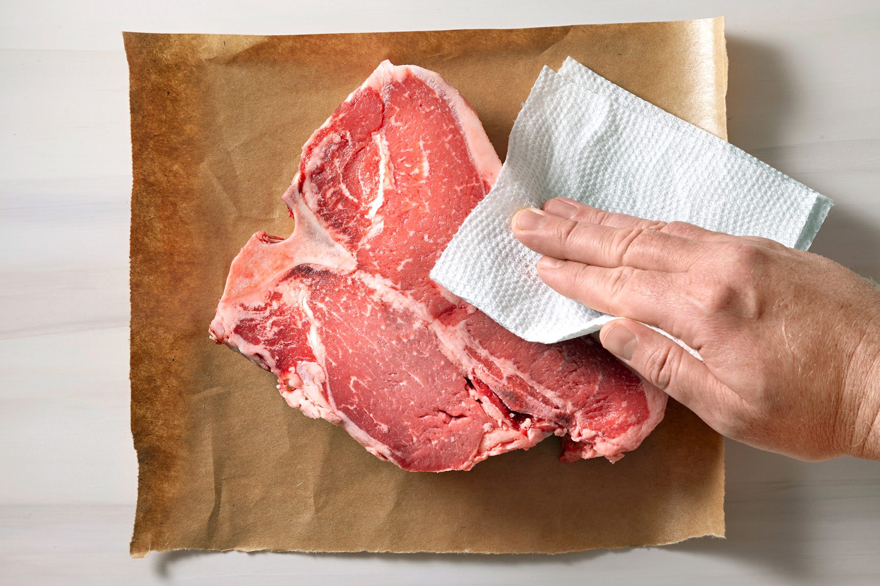 A steak is being patted dry with a paper towel to remove excess moisture.